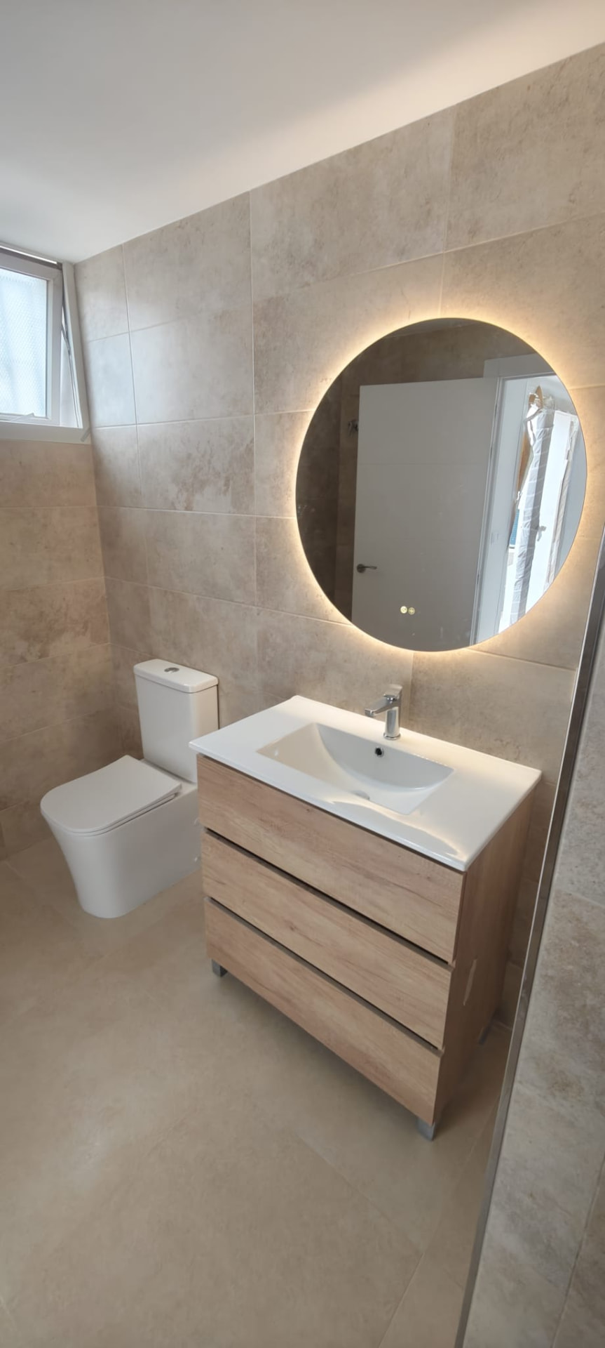 Aseo con revestimiento tipo travertino, espejo circular retroiluminado y mueble de madera