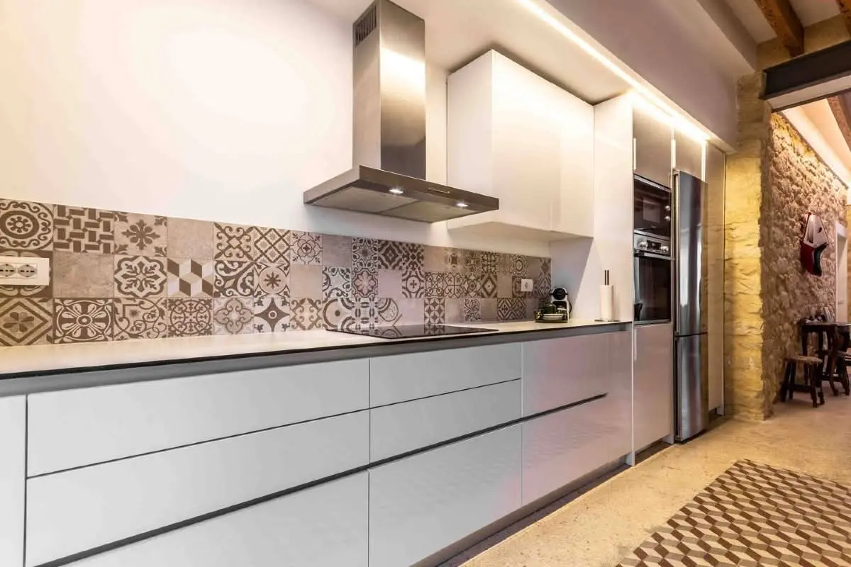 Cocina lineal con frentes lacados, campana de acero y frente de baldosa hidráulica junto a muro de piedra