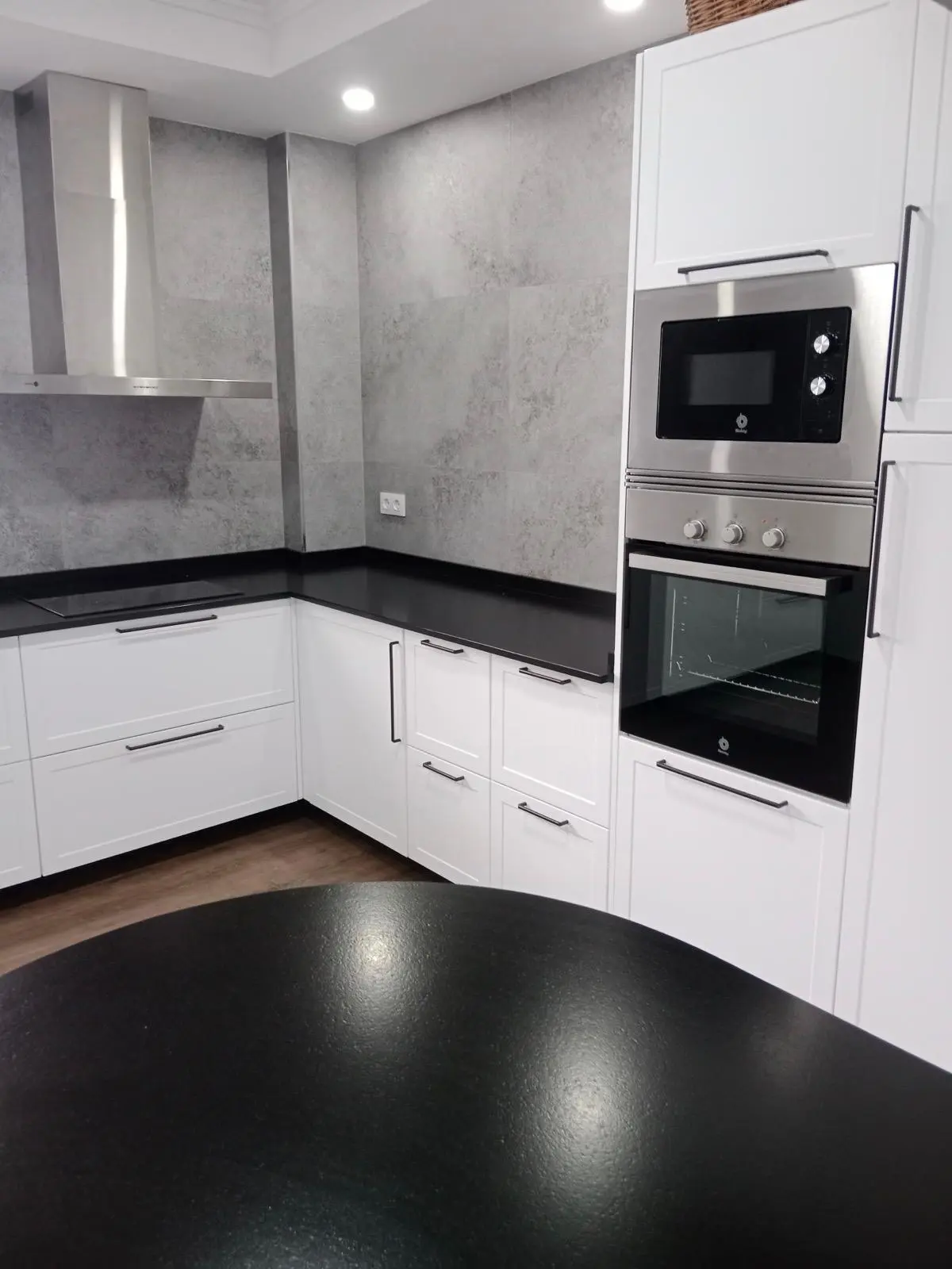 Cocina en L con muebles blancos, encimera negra y revestimiento tipo cemento